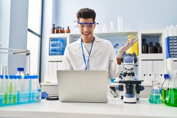 Wall Mural - Young hispanic man working at scientist laboratory doing video call celebrating achievement with happy smile and winner expression with raised hand