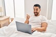 © Krakenimages.com - Young hispanic man with beard using computer laptop on the bed pointing thumb up to the side smiling happy with open mouth