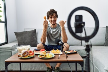 Sticker - Young hispanic man recording eating food video with smartphone at home pointing thumb up to the side smiling happy with open mouth