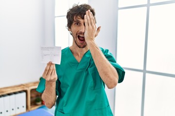 Sticker - Young hispanic doctor man holding covid record card covering one eye with hand, confident smile on face and surprise emotion.