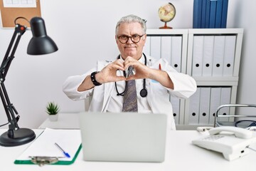 Canvas Print - Senior caucasian man wearing doctor uniform and stethoscope at the clinic smiling in love doing heart symbol shape with hands. romantic concept.