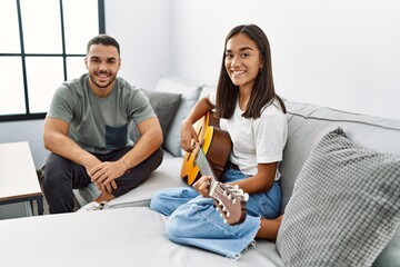 Sticker - Young latin couple playing classical guitar sitting on the sofa at home.