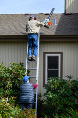 Wall Mural - Senior couple working on fall maintenance, woman holding extension ladder while man climbs up with a gas powered leaf blower
