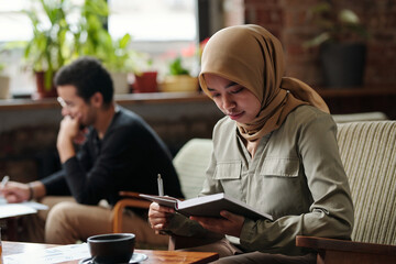 Aufkleber - Young serious Muslim businesswoman in hijab holding open notebook in front of herself and looking through notes by workplace