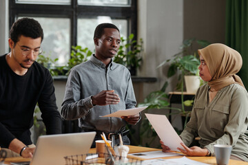 Aufkleber - Two young intercultural colleagues discussing financial documents by workplace while multi-ethnic male analyst standing in front of laptop