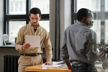Aufkleber - Young serious businessman looking through financial document while standing by table in front of African American male colleague