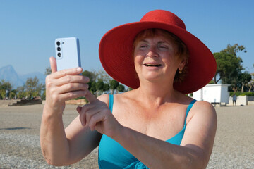 Wall Mural - Senior adventureisageless woman wearing in swimsuit and red hat taking a selfie on a mobile phone on the beach. Summer vacation for pensioner. Internet for holiday