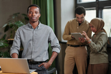 Poster - Young confident business leader in smart casualwear standing by workplace in front of camera against two colleagues