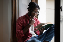 Young Focused Black Man Wearing Eyeglasses Learning Foreign Language By Reading Books, African American Student Guy Sitting On Windowsill At Home Learning From Textbook. College Exams Preparation