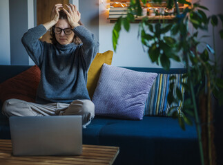 Upset young woman holding her head while sitting on sofa in front of laptop, headaches and burnout at online work