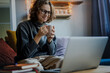 © olezzo - Young smiling woman in eyeglasses and warm sweater sitting on a sofa in front of a laptop with a mug of coffee and looking at the screen