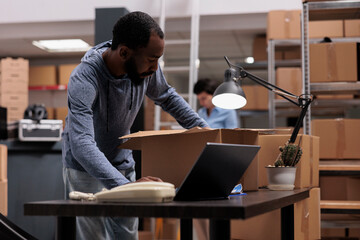 Worker putting client order in cardboard box wrapping with bubble wrap for protection, checking shipping details on laptop computer before delivery package. Employee working in distribution center