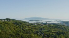 A Wonderful Morning View Of The Mountain Village With The Misty Haze And Fog Shrouding Distant Mountains In The Background, Surrounded By Tall Trees And Green Fields.