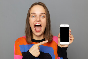 Portrait of happy amazed woman with brown hair wearing colorful jumper, pointing at smart phone with smart phone with empty display, keeps mouth open, posing isolated over gray background.