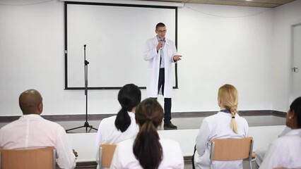 Canvas Print - Confident male speaker in white coat giving presentation from stage at medical conference. High quality FullHD footage