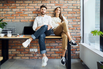 Smiling young entrepreneurs business partners sitting together on the desk in small startup office