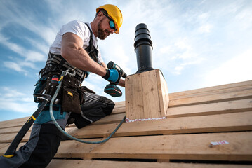 a carpenter with a safety harness and tool belt holds a electric screwdriver in his hand. he is work