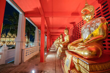 Several Golden Statues Of Buddha Statues In Meditation Posture Are Lined Up In A Corridor And Spotlight On Every Statues In Front Of The Red Wall.