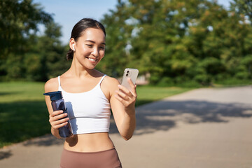 Fitness woman with water bottle and smartphone, jogging in park and smiling, looking at her mobile phone app, checking sport application