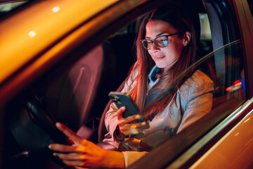 Business woman driving a car in a city during a night and using smartphone