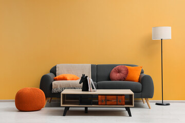 Stylish grey sofa with colorful pillows, wooden table and lamp near pale orange wall indoors. Interior design