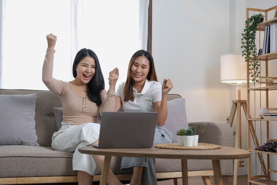 Two happy Asian women best friends in casual wear laughing while working with laptop at home in living room