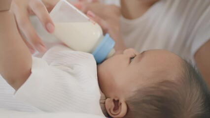 Wall Mural - Closeup view of Young asian mother feeding her little cute baby daughter with bottle on bed.Asian young mother See also baby and asian newborn baby suck milk from bottle and sleep on white bed