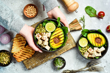 Wall Mural - Female hands holding a bowl of vegan lunch: avocado, tofu cheese, flax, pumpkin seeds and spinach. The concept of healthy eating. Top view.