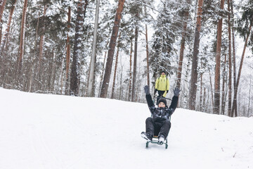 Wall Mural - Father with backpack and little sos walking together in winter snowy forest. Happy man and joyful boy sledding and having fun together. Wintertime activity outdoors. Concept of local travel