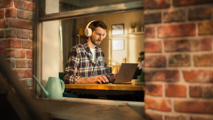 Aufkleber - Handsome Caucasian Man Working on Laptop Computer while Sitting Behind His Desk at Home Office. Freelance Entrepreneur Listening to Music, Doing Remote Telework. View From Outdoors into Window.