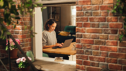 Poster - Hispanic Woman Using Laptop Computer and Sitting in Living Room im Her Apartment. Professional Female Working Remotely Online, Planning, Brainstorming New Project. Shot Into the Window From Outside.
