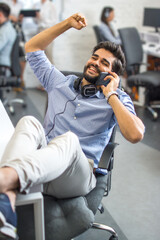 Wall Mural - Exited young business manager talking on phone while having break at office, leaning back on chair and putting legs on table at his workspace