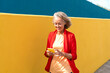 © VISTA by Westend61 - Smiling mature woman using mobile phone walking by yellow wall