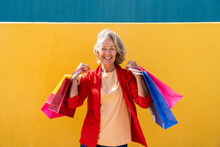 Happy Mature Woman With Multi Colored Shopping Bags In Front Of Yellow Wall