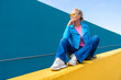 © VISTA by Westend61 - Mature woman wearing blue leather jacket sitting on yellow wall