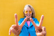 © VISTA by Westend61 - Happy mature woman gesturing peace sign in front on yellow wall