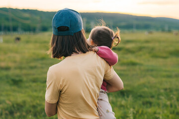 Wall Mural - Back view of a mother wearing hat carrying the baby child in her arms, walking through the green field. Carefree childhood. People lifestyle. Summer vacation