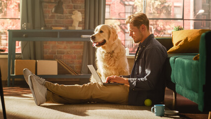 Fototapete - Portrait of a Handsome Young Adult Male Sitting on a Floor and Using Laptop Computer in Sunny Loft Living Room. Golden Retriever Dog Sitting Next to Him and Wanting to Play and Get Petted.
