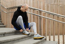A Young Male Athlete Sits On The Steps And Ties His Shoelaces Before A Running Workout. A Bald Man With Freckles Listen Music On Wireless Headphones And Prepares For A Training.