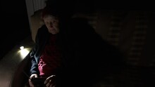Ukraine. Grandmother Holds Radio In Hands Sitting In Darkness Blackout During Russian Terroristic Attacks. Senior Woman In Winter Clothes In Darkness On Sofa With Hand Radio Receiver Listening News.