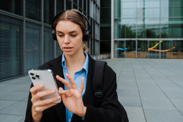 Young business woman using headphones and mobile phone while walking at city street