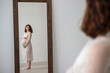 © Michael - a pregnant brunette girl in a white wedding dress with a big belly straightens looking into a mirror and her reflection