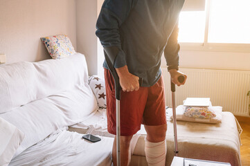 Injured man with bandaged foot walking at home with walking sticks.