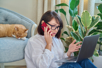 Wall Mural - Mature woman sitting at home, using smartphone laptop