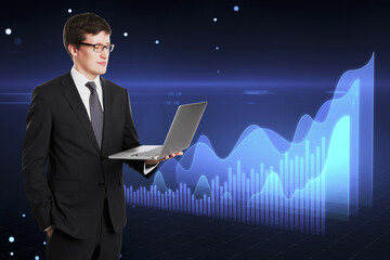 Poster - Attractive young european businessman holding laptop with glowing blue business chart hologram on blurry dark tech background. Price, exchange, online banking and money concept.