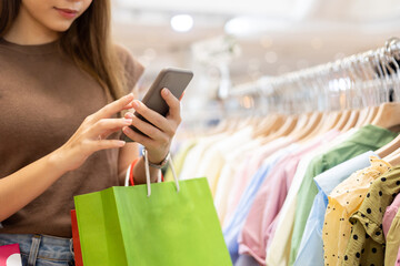 happiness young beautiful woman with shopping bag using cell phone choosing dress at row of clothes 