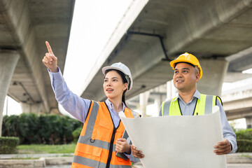 Wall Mural - Two Asian worker engineer man and woman architect looking construction with white safety helmet in construction site. Standing at highway concrete road site. Work planning with blueprint and tablet.