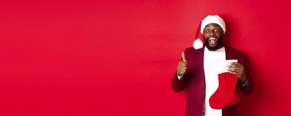 Excited Black man showing thumb-up in approval, holding christmas sock with holiday gifts, smiling amazed, standing over red background