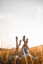 Woman And Man Having Fun Outdoors. Loving Hipster Couple Are Lying In The Grass And Lifting Their Legs In Sneakers Up In The Summer At Sunset Light. Valentines Day.