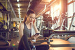 Beautiful young sporty woman with earphones using phone and listening to music during the break at gym. Fitness woman using phone in gym with towel around neck.